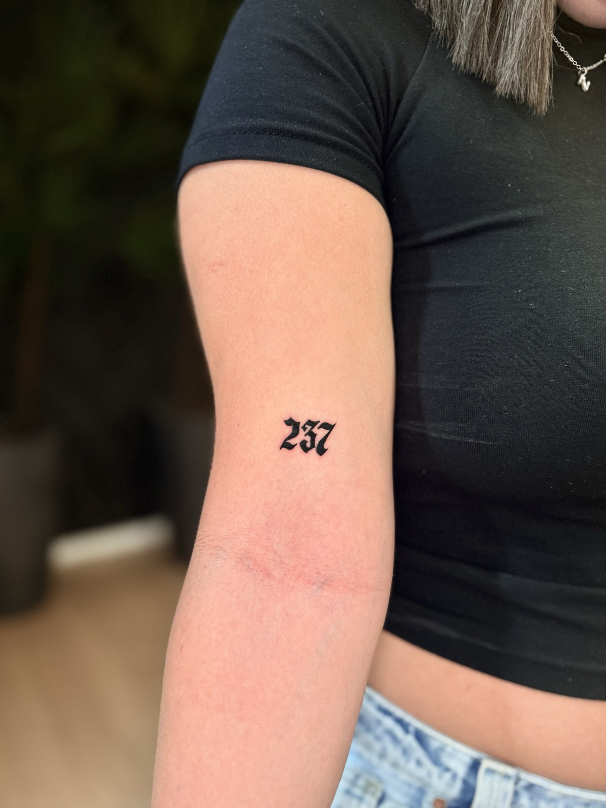 Número 237 em letra gótica preenchida de preto, na parte de trás do braço. Por Dudis (Maria Eduarda Freitas), na ADV Tattoo.