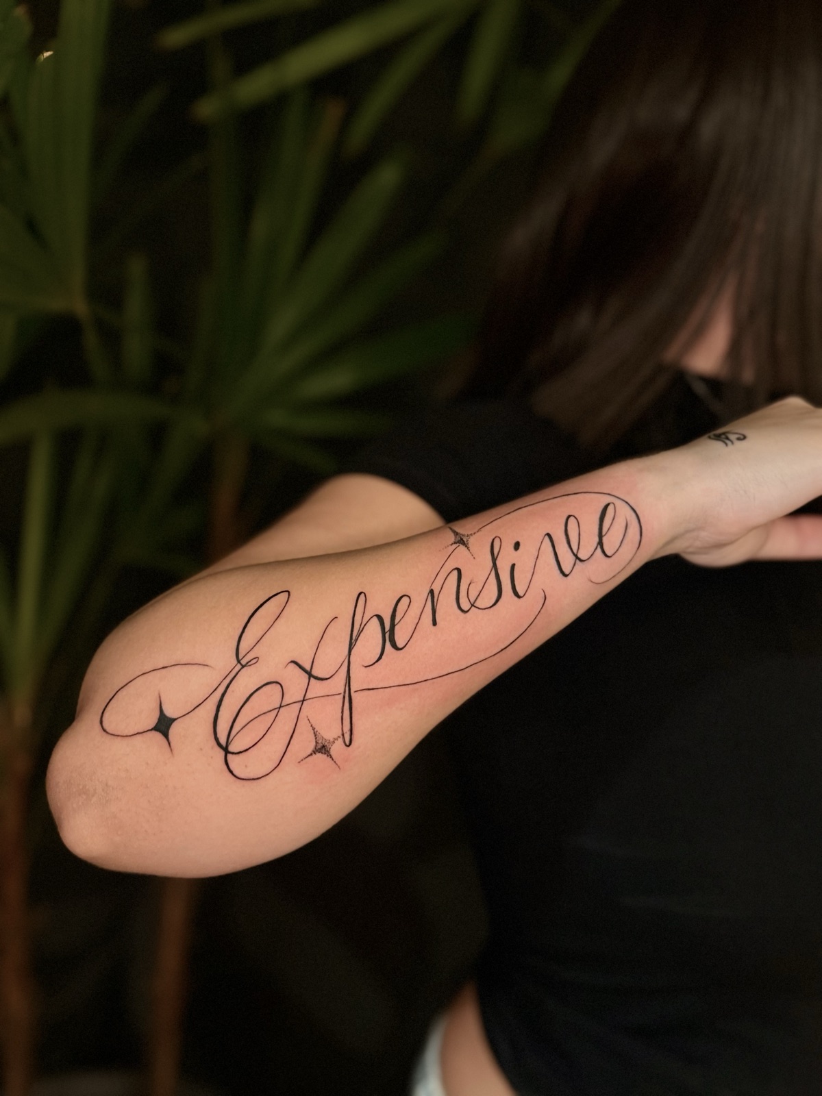 Palavra "Expensive" em caligrafia com floreios longos e estrelas em pontilhismo, ao longo do antebraço. Por Dudis (Maria Eduarda Freitas), na ADV Tattoo.